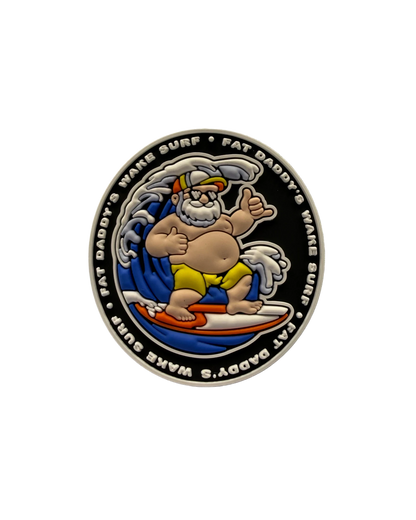 A circular enamel coin or medallion featuring a cartoon surfer character on a white surfboard against a black background, with the text 'FAT DADDY'S WAVE SURF' around the edge.