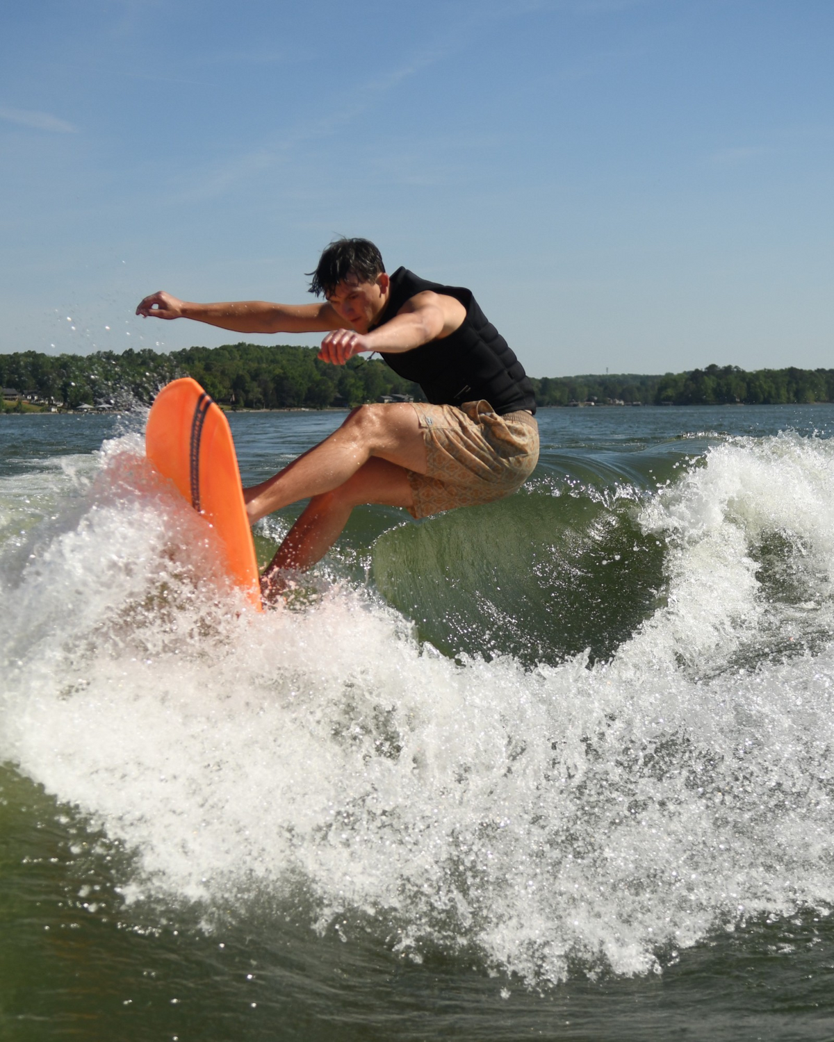 Man riding a bright orange Barrette Greenroom wakesurf board on a lake wave while wearing a black life vest